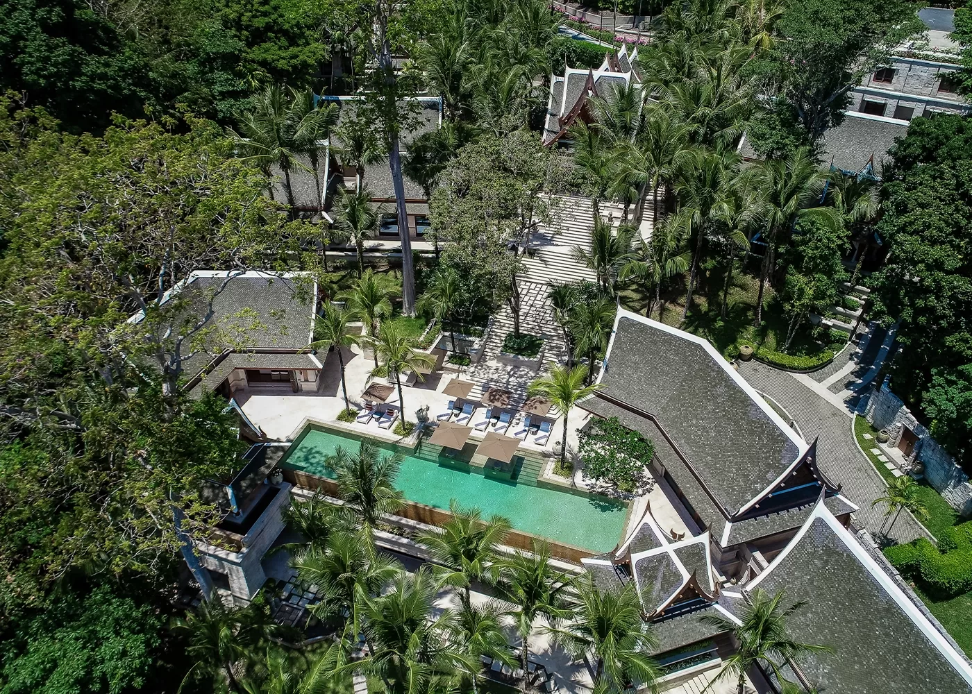 Aerial view of a tropical resort with a rectangular swimming pool surrounded by lounge chairs, palm trees, and several traditional-style buildings with pitched roofs, set amidst lush greenery.