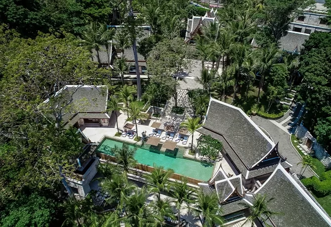 Aerial view of a tropical resort with a rectangular swimming pool, lounge chairs, palm trees, and traditional-style buildings surrounded by lush greenery.