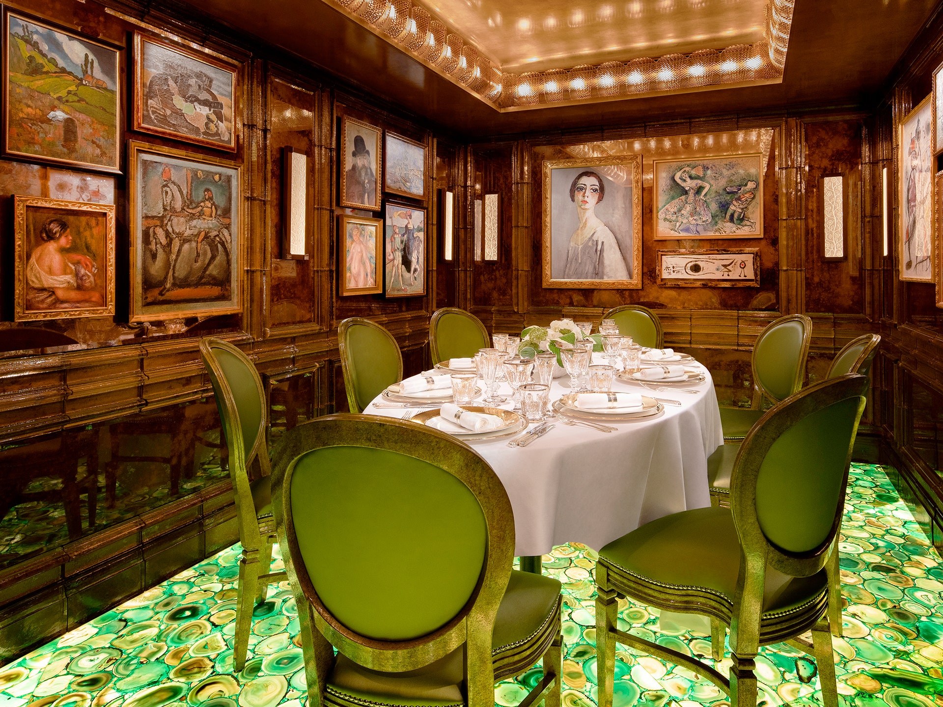 Private round dining table and green luxury dining chairs in wooden room filled with framed paintings on abstract green backlit gemstone floor