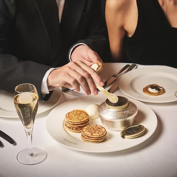 Two elegantly dressed people sit at a white-clothed table, serving caviar and cream onto blinis. A glass of champagne, stacked blinis, and an open tin of caviar are arranged on a white plate in front of them.