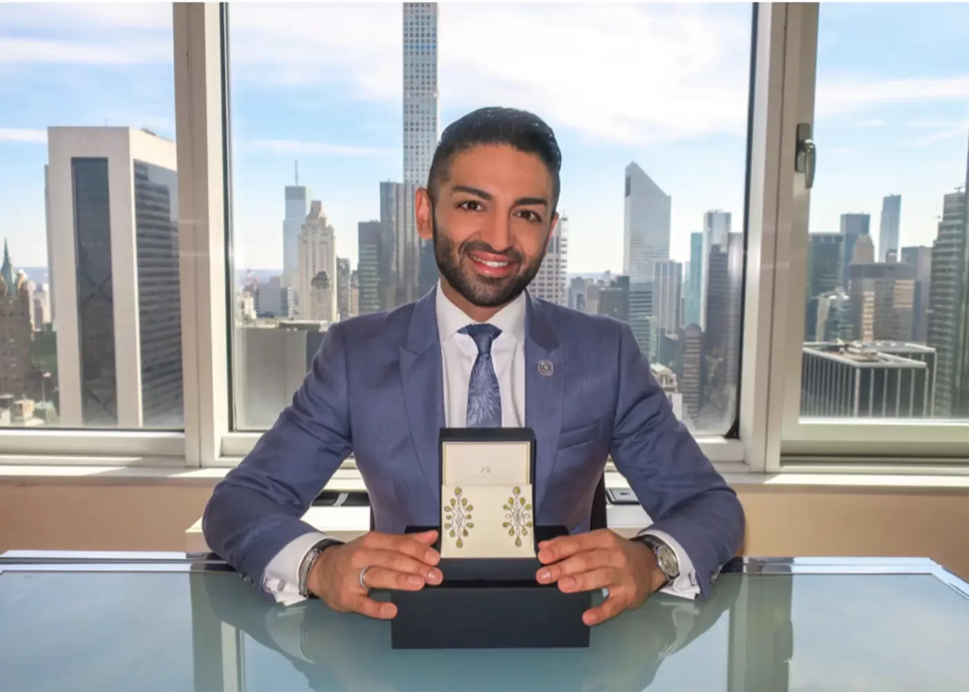 A man in a blue suit sits at a glass desk in front of large windows with a city skyline view, smiling and holding an open jewelry box displaying a pair of gold earrings.