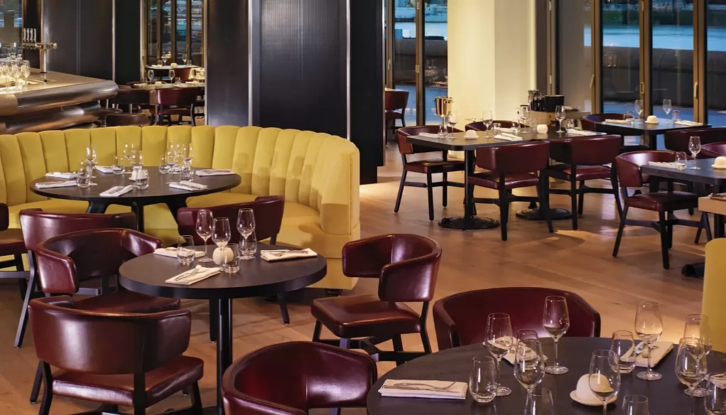 Elegant restaurant interior with a mix of yellow booth seating and dark wooden chairs around round tables. Place settings include wine glasses and cloth napkins. Large windows in the background offer a view outside at Sea Containers, London.