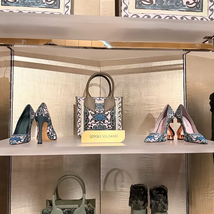 A display shelf shows a patterned handbag and two pairs of matching high-heeled shoes with blue, white, and black designs. A sign with the brand name Sergio Soldano is placed in front of the handbag.
