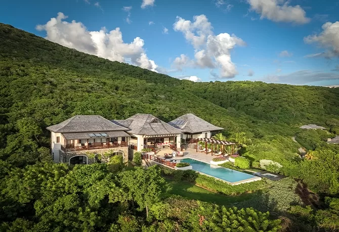 Aerial view of a luxurious villa with a swimming pool nestled in lush green hills under a blue sky with scattered clouds.