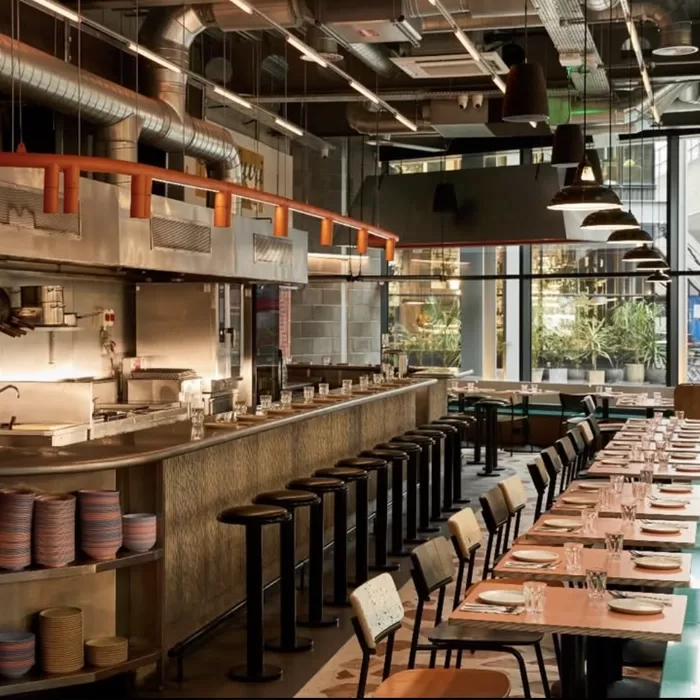 A modern restaurant interior with an open kitchen, high bar stools, and a long dining table set with plates and glasses. Large windows let in natural light, and industrial-style lighting hangs from the ceiling.