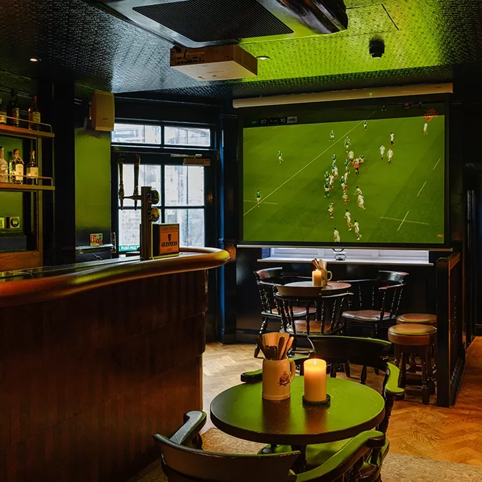 A cosy pub interior with wooden tables and chairs, a bar counter, and a lit candle on a table. A large screen on the wall shows a rugby match in progress. The atmosphere is warm and inviting.