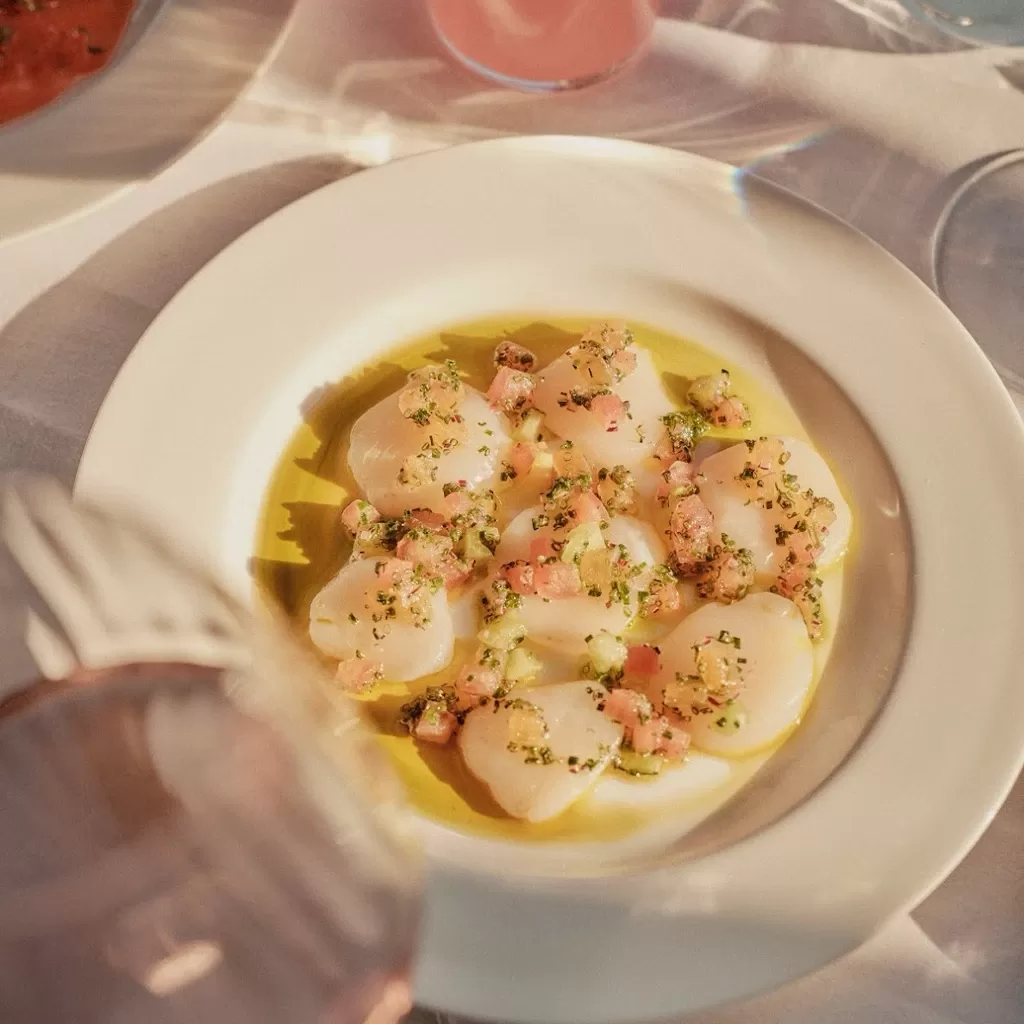 A white plate with scallops drizzled with olive oil and topped with finely diced herbs and vegetables, set on a sunlit table with a pink drink and shadows visible nearby.