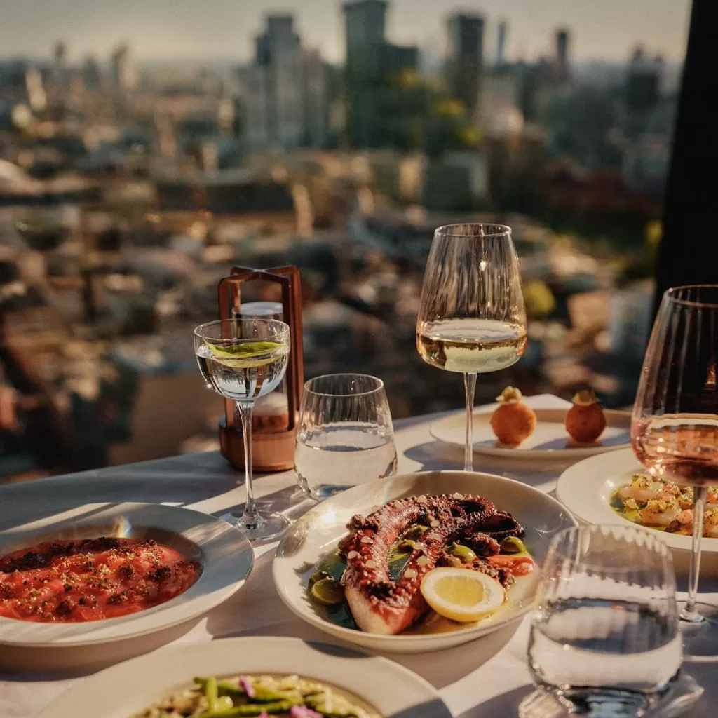 A table set with gourmet dishes, wine glasses, and water glasses overlooks a cityscape through a large window on a sunny day. Elegant plates of seafood, pasta, and appetizers are displayed on a white tablecloth.