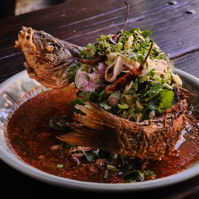 A whole fried fish served upright in a shallow bowl, topped with a vibrant mix of fresh herbs, onions, and red chilies, surrounded by a rich, dark sauce on a rustic wooden table.