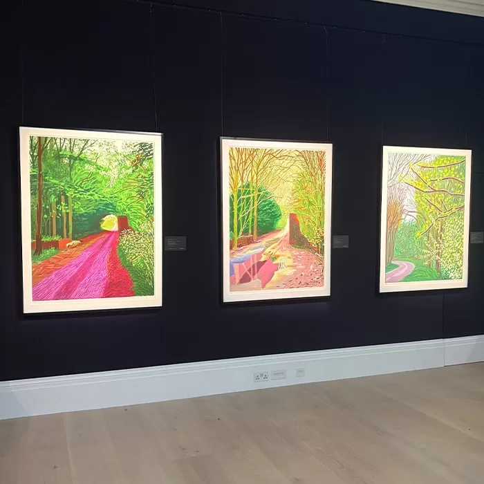 Three brightly colored landscape paintings hang on a dark wall in an art gallery. Each features a different path or road winding through vivid, stylized trees with bold, contrasting hues.
