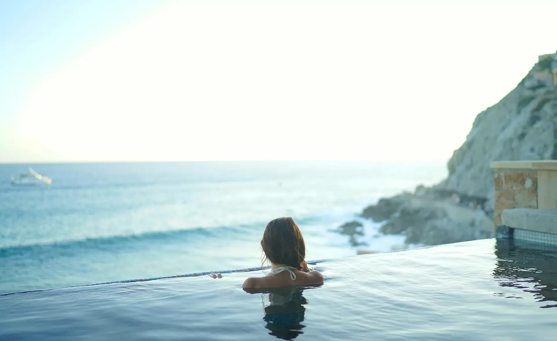 A person in a swimsuit leans on the edge of an infinity pool, overlooking a serene ocean view. Waves gently meet the rocky shore, and the sky is bright and clear, suggesting a calm and peaceful atmosphere.