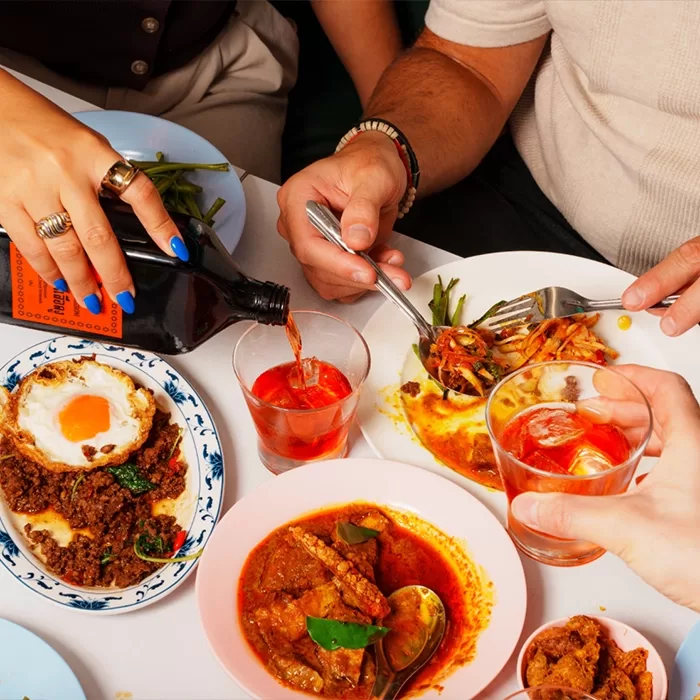 Three people share a meal at a table with vibrant dishes, including curry, eggs, and vegetables. One hand pours a dark drink into a glass while others reach for food with forks and hold drinks.