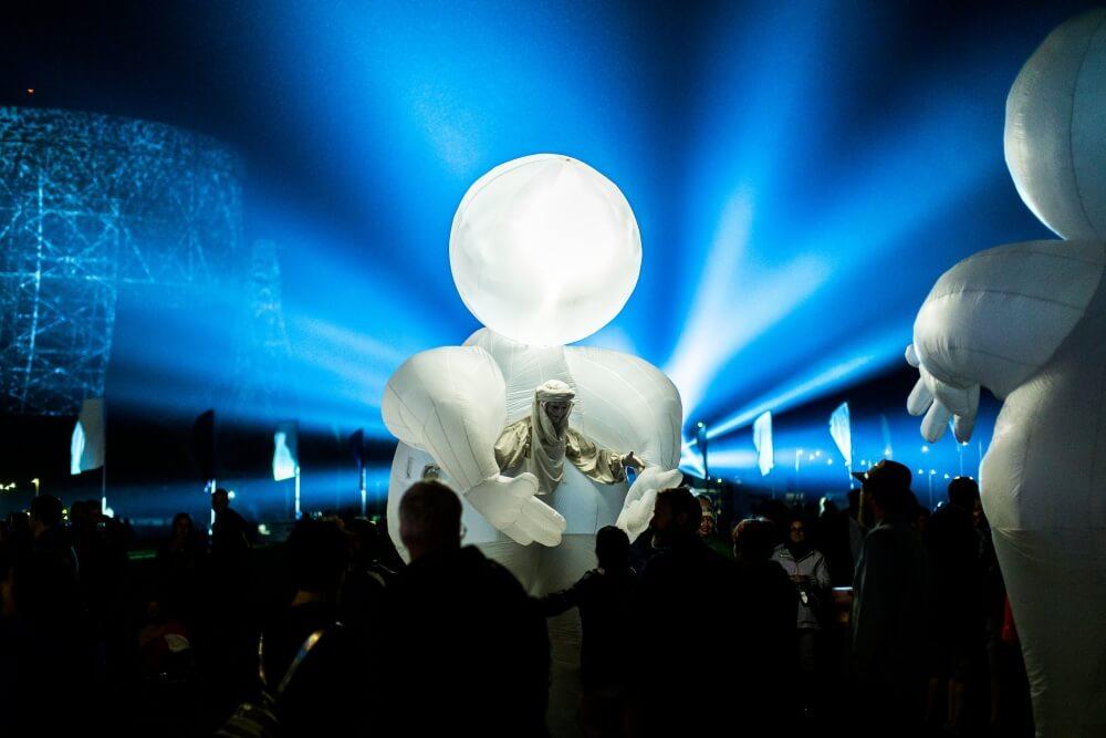 Huge white blown up figure with hands with a person stood between amongst a crowd at a stage with blue lights at Bluedot.