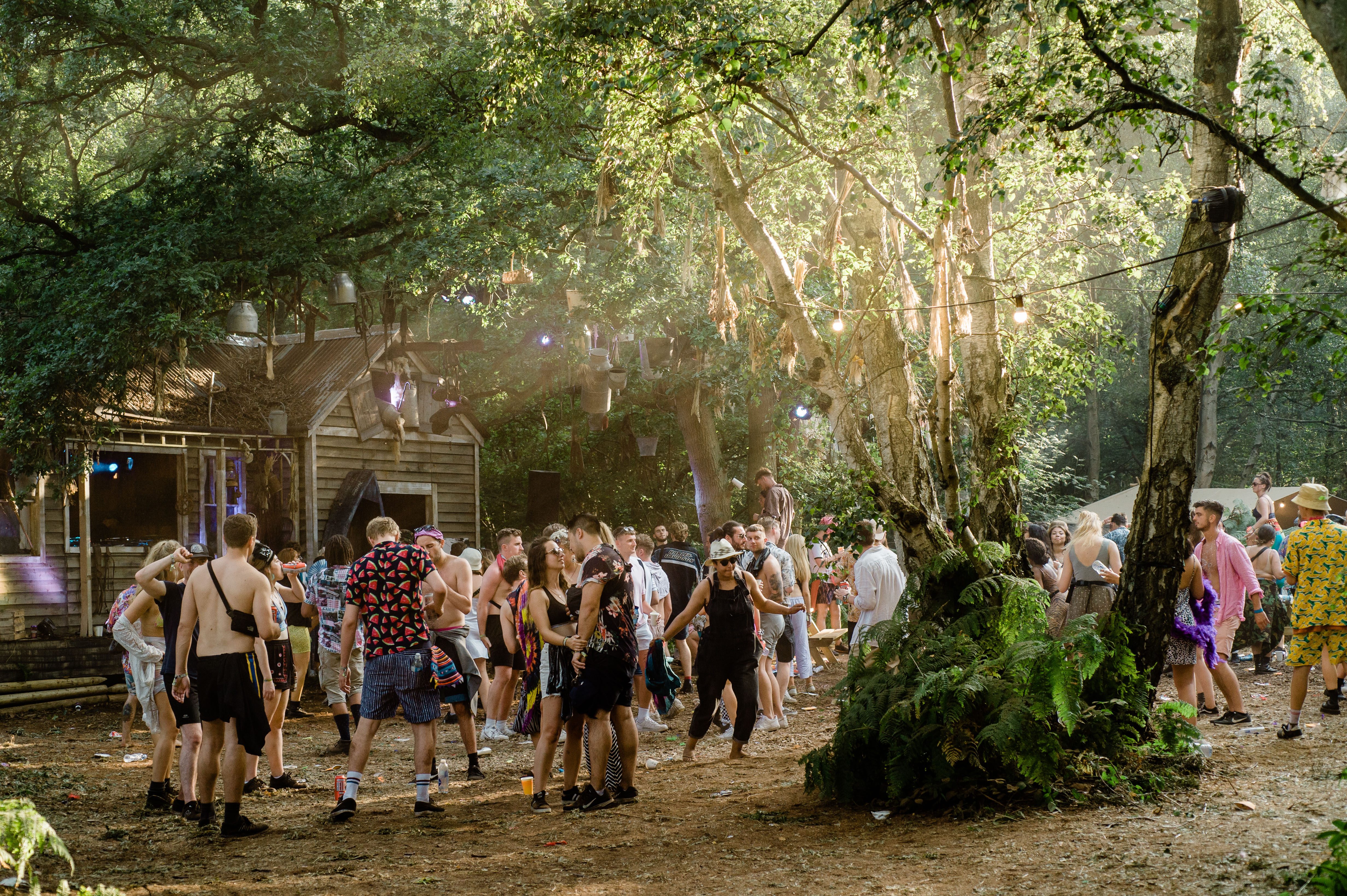Outside stage at Lost Village with a crowd of young adults dressed in funky colourful festival clothing dance together between the trees to the dj in an outside hut.
