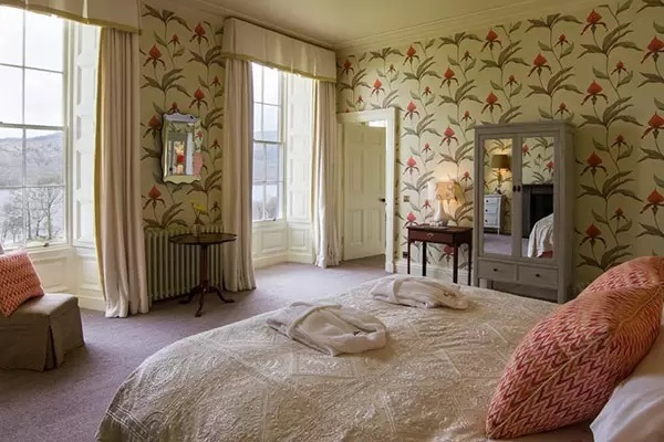 Inside hotel room white double bed with red pillows, mirrored wardrobe, printed wallpaper and tall glass windows.