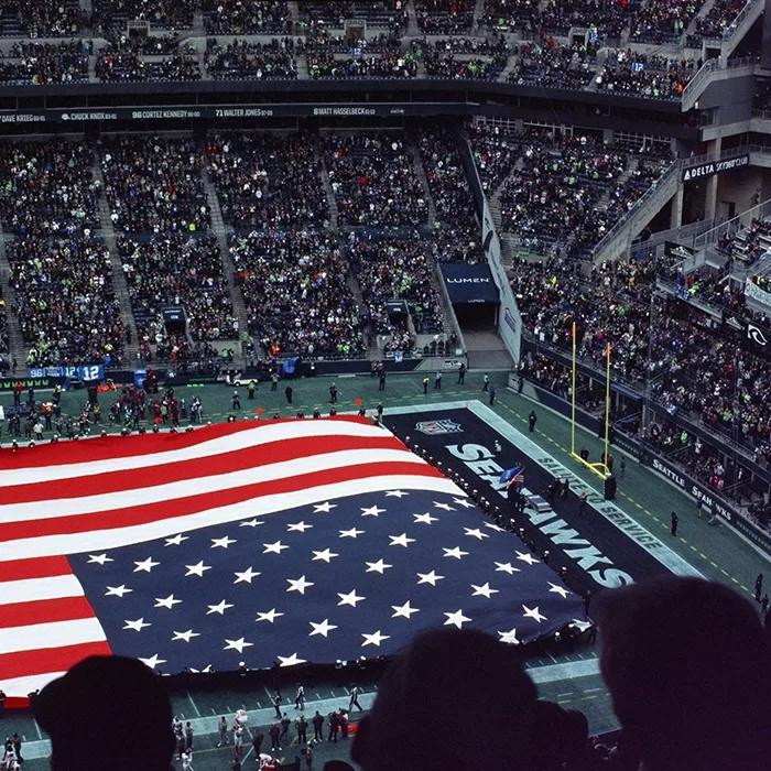 A large American flag is held across the field at a football stadium filled with fans. The stadium displays “Seahawks” branding, and the crowd watches as the flag is presented before the game.