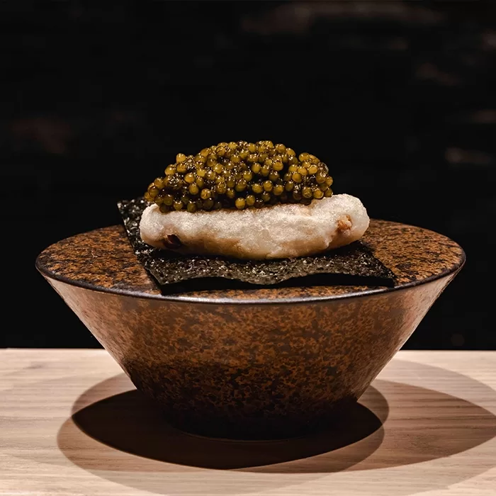 A ceramic bowl holds a crisp sheet of seaweed topped with a tempura-fried piece and a generous layer of golden caviar, set against a dark background.