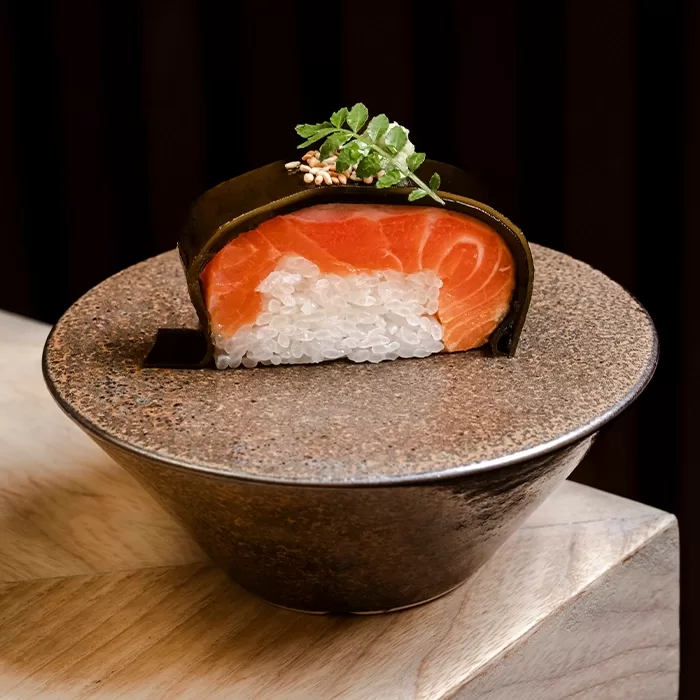 A beautifully plated sushi piece with a thick slice of salmon on top of compact white rice, wrapped in seaweed, and garnished with microgreens and sesame seeds, served in a rustic bowl.