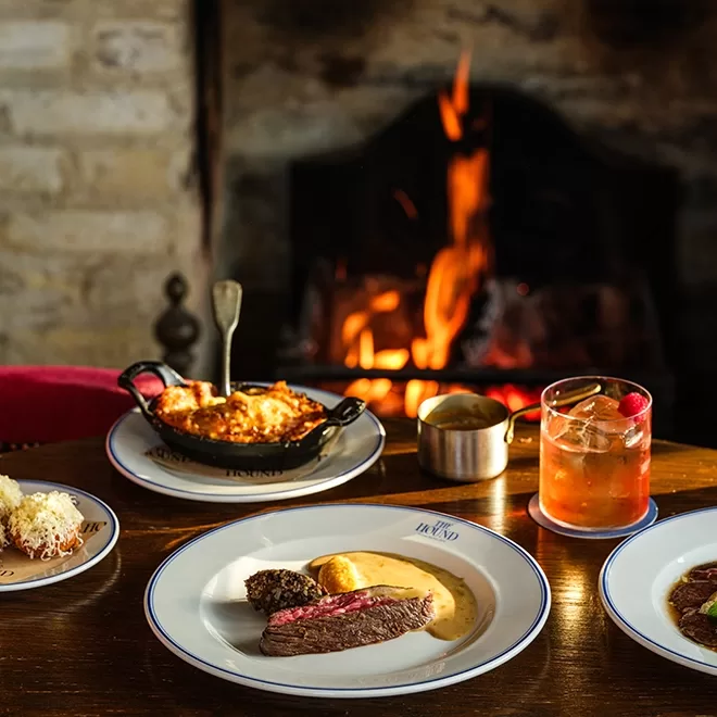 Plates of gourmet food, including steak with sauce and a casserole, are set on a wooden table in front of a fireplace. A cocktail with ice and fruit sits next to the dishes, creating a cozy dining atmosphere.