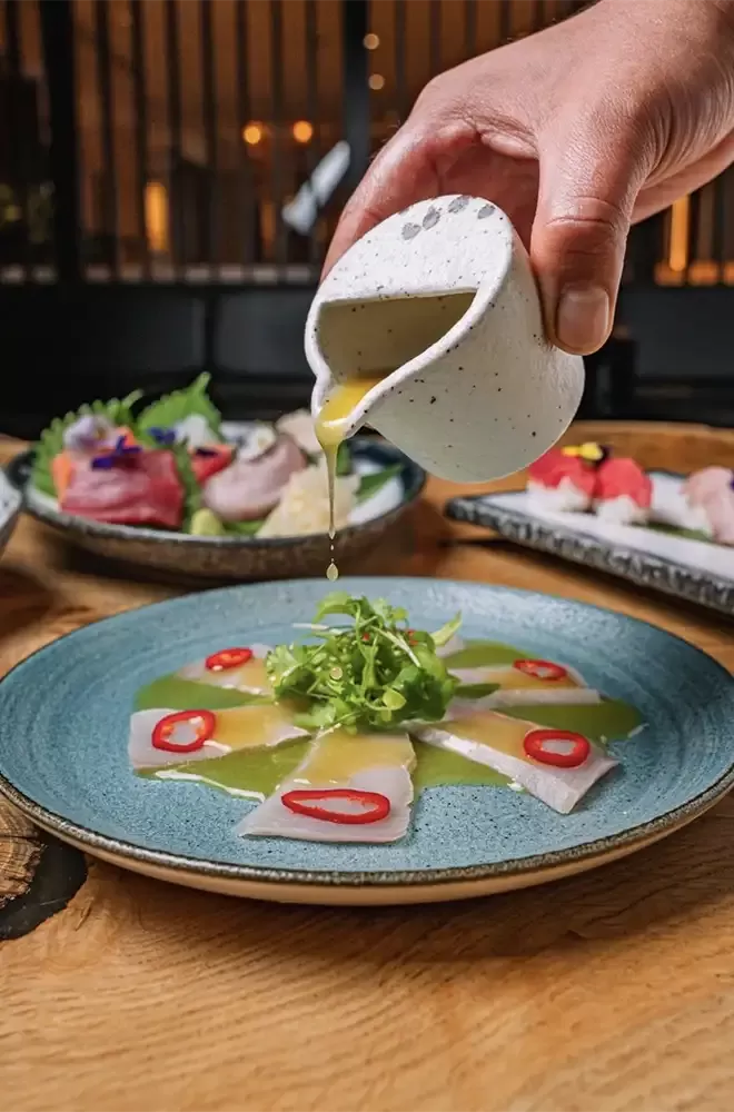 A person elegantly pours sauce over a beautifully plated dish at Tokii restaurant, enhancing the meal's presentation.