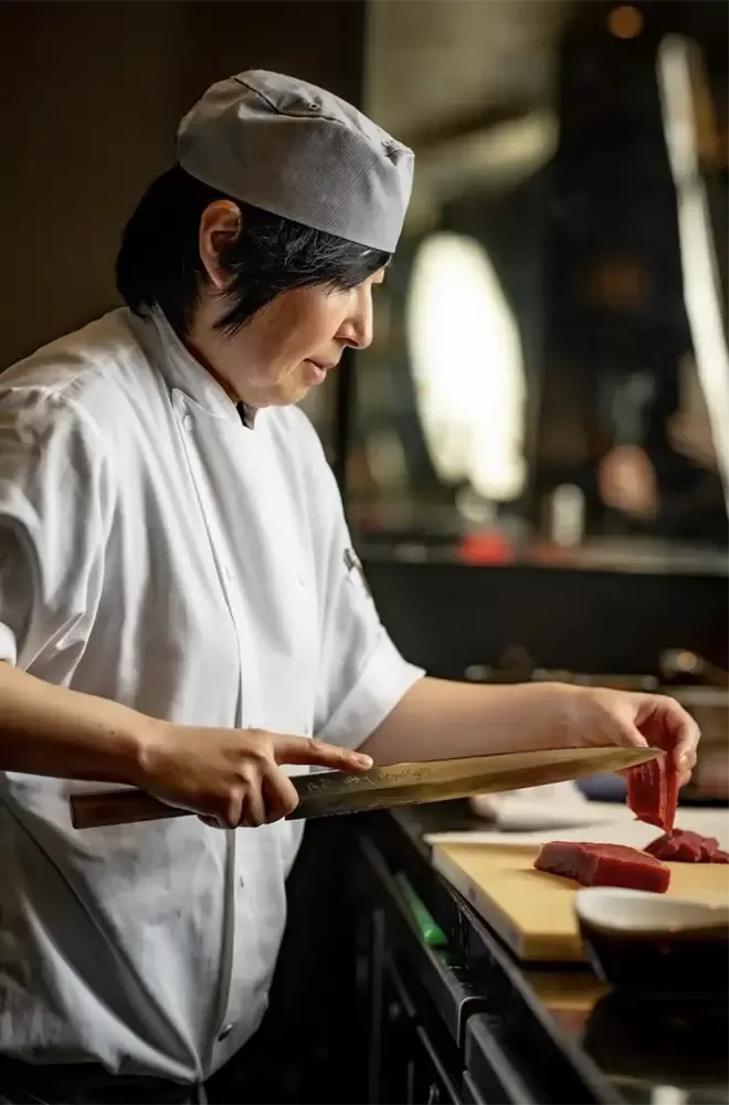 A woman in a chef hat skillfully prepares food at Tokii restaurant, showcasing her culinary expertise.