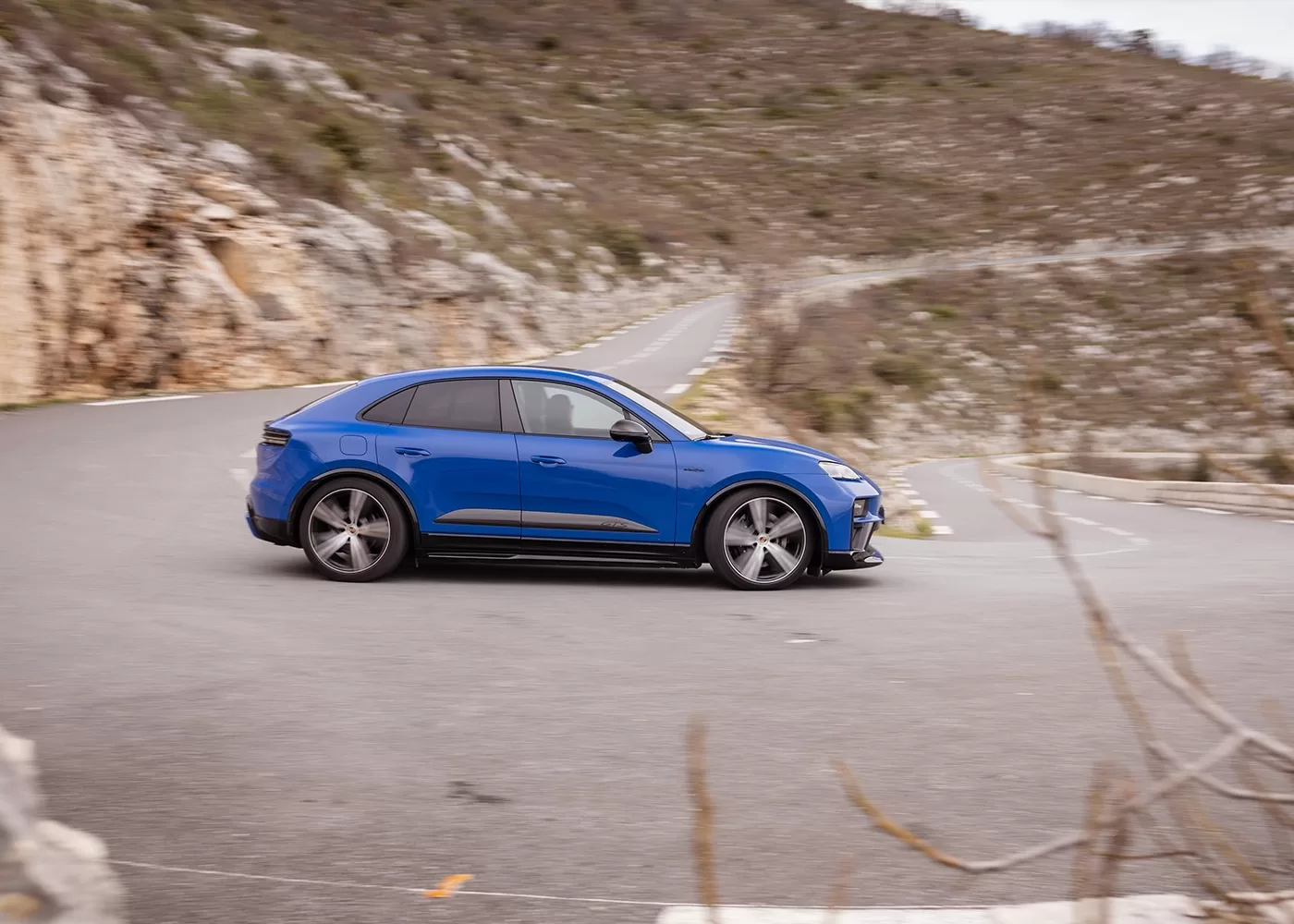 A blue sedan-style car drives around a sharp curve on a winding mountain road, surrounded by rocky hills and sparse vegetation.