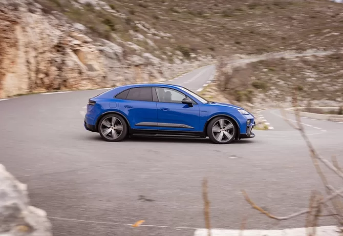 A blue car is driving around a sharp curve on a winding mountain road, with rocky hillsides and sparse vegetation in the background.
