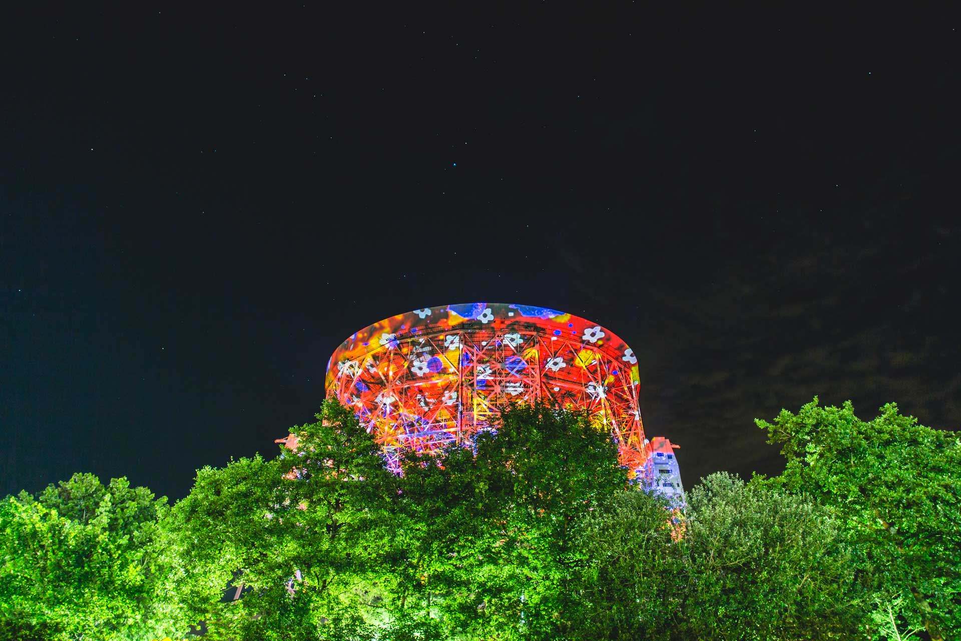 Worms-eye view of tall round colourful stage which is covered in red, blue, yellow lights and white flowers in between tall green trees.