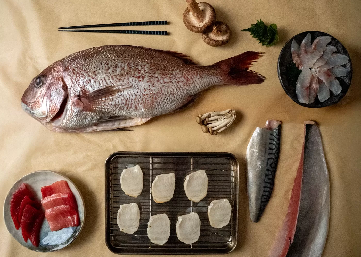 A flat lay of fresh fish and ingredients for sashimi: a whole fish, sliced raw fish, mushrooms, green herbs, chopsticks, and scallops on a tray, arranged neatly on a beige surface.