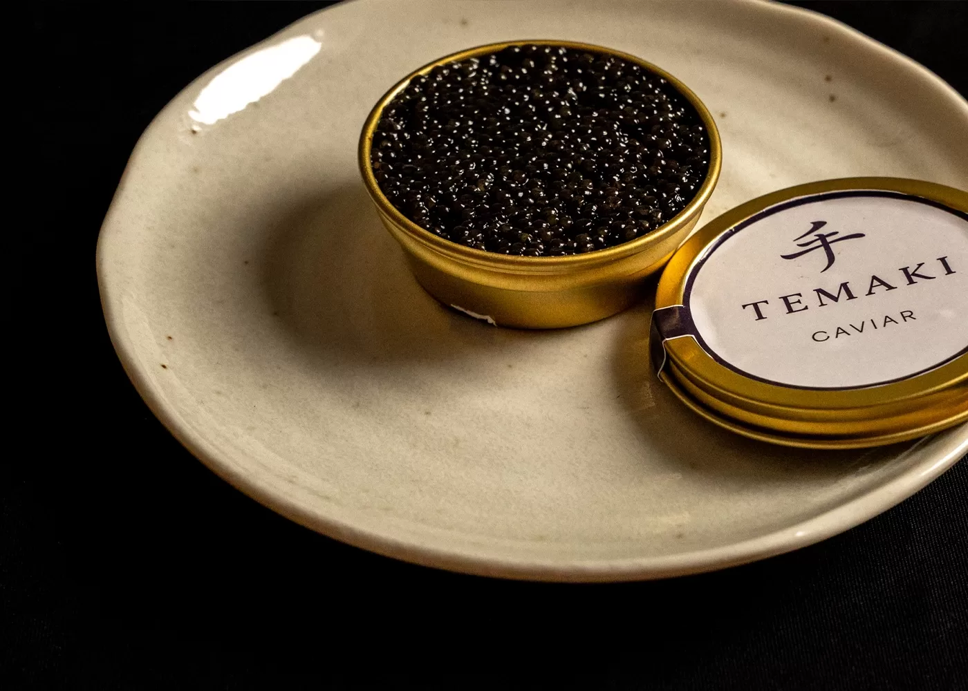A small gold tin of black caviar with TEMAKI CAVIAR labeled on the lid sits on a plain white plate against a black background.