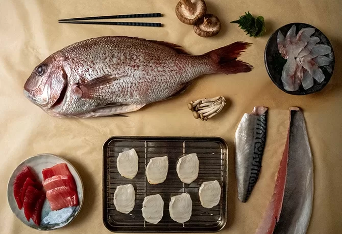 A whole fish, fish fillets, sliced raw fish, scallops, mushrooms, leafy greens, and chopsticks are arranged on a beige surface, showcasing ingredients for Japanese cuisine or sashimi preparation.