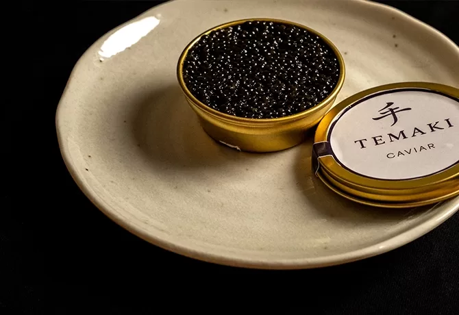A small tin of black caviar, labeled TEMAKI Caviar, sits open on a plain white plate against a dark background. The lid is placed beside the tin on the plate.