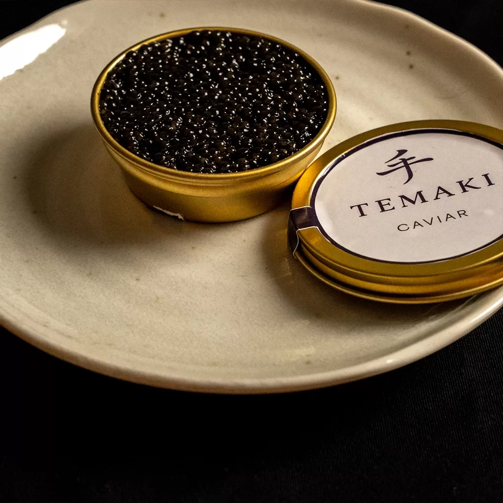 A tin of black caviar labeled TEMAKI Caviar sits open on a white plate, with the lid placed beside it on a dark surface.