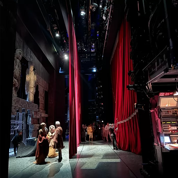 Backstage view of a theater with people in costumes standing near red curtains. Stage props and set pieces are visible, along with technical equipment and lighting.
