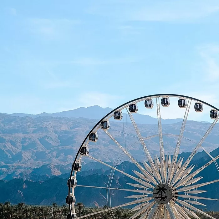 A Ferris wheel with enclosed gondolas stands in front of distant mountain ranges under a clear blue sky.