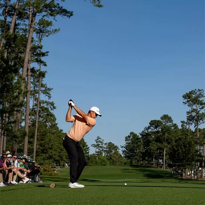A golfer in a peach shirt and white cap prepares to swing on a lush, green golf course lined with tall trees, while a group of spectators watches from the side under a clear blue sky.