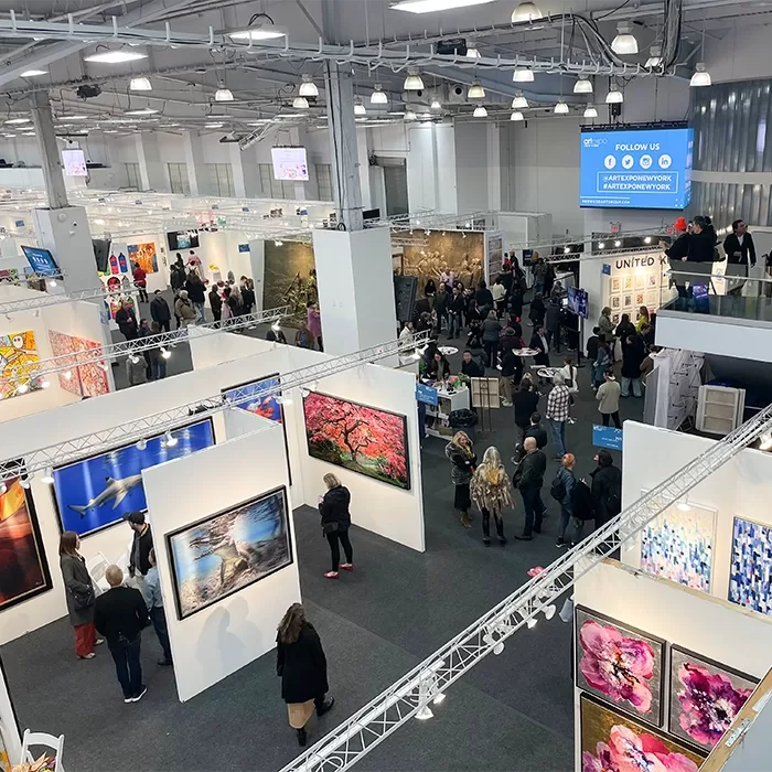 A busy indoor art fair with people walking and viewing colorful paintings displayed on white partition walls under bright lights in a spacious exhibition hall.