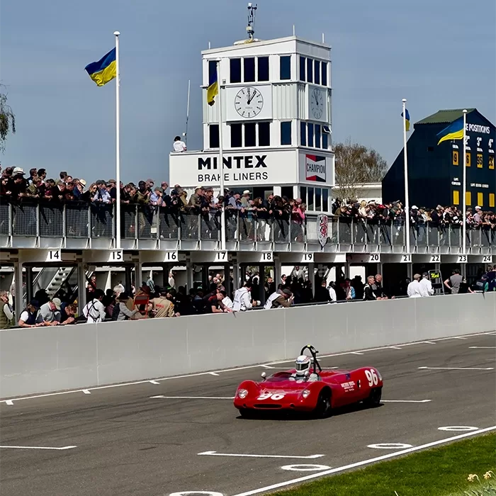 A red vintage race car numbered 96 drives past a crowd of spectators and a race control tower with a large clock at a classic car event on a sunny day.