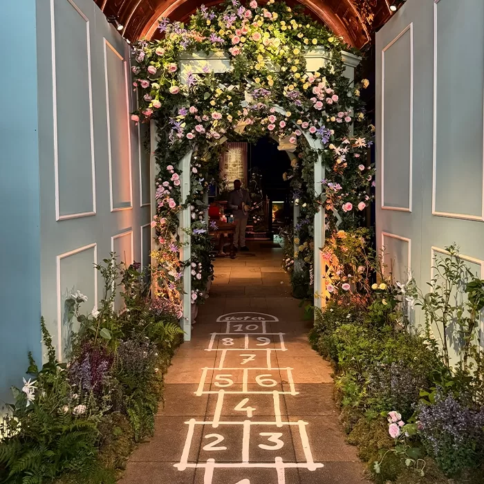 A hopscotch game is painted on a stone path leading through an archway covered with flowers and greenery, inside a space decorated with pastel blue walls and more flowers.