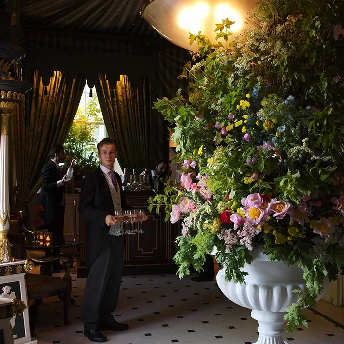 A formally dressed waiter stands beside a large, ornate white vase filled with lush, colorful flowers in an elegant, dimly lit room with green drapes and a bar in the background.