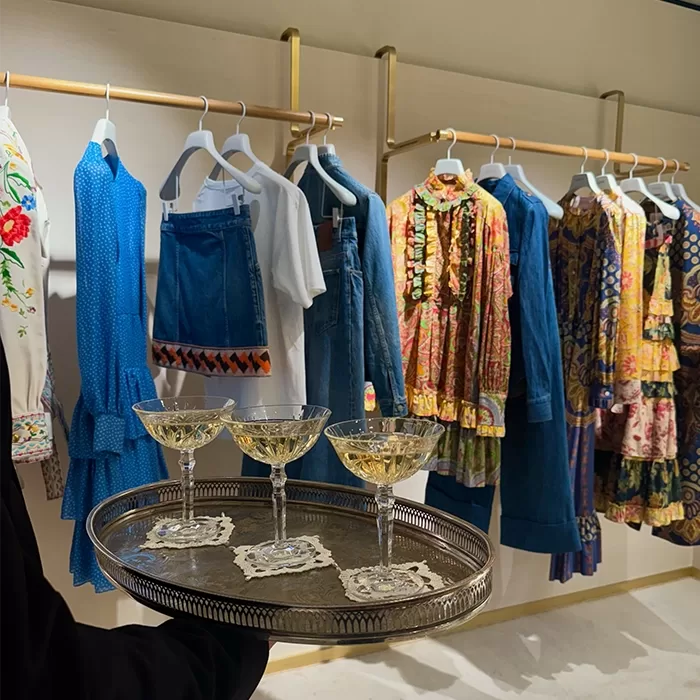 A person holds a silver tray with three filled champagne glasses in front of a clothing rack displaying colorful and denim garments on white hangers.