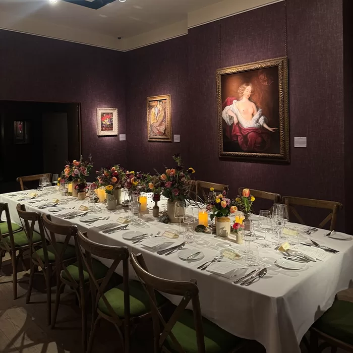 A long, elegant dining table is set with candles, floral centerpieces, and place settings in a dimly lit room with dark purple walls, wooden chairs with green cushions, and classical paintings hung on the walls.