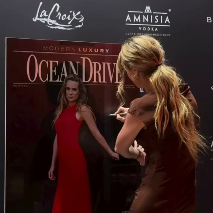 A woman with long, wavy hair signs a large Ocean Drive magazine cover featuring another woman in a red dress at a black backdrop event featuring LaCroix and Amnesia Vodka logos.