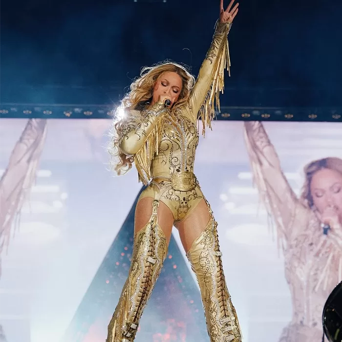 A performer in a gold, fringe and sequin outfit sings on stage with one arm raised. Large images of the performer are displayed on screens in the background.