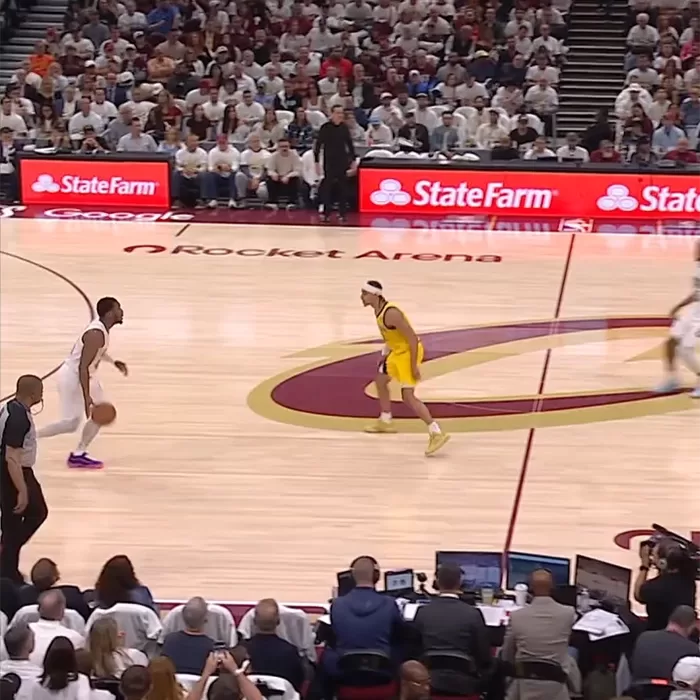 Two basketball players face each other on a court during a game; one in a white uniform dribbles the ball near half-court, while another in yellow defends. The arena is full of spectators, with Rocket Arena visible on the floor.