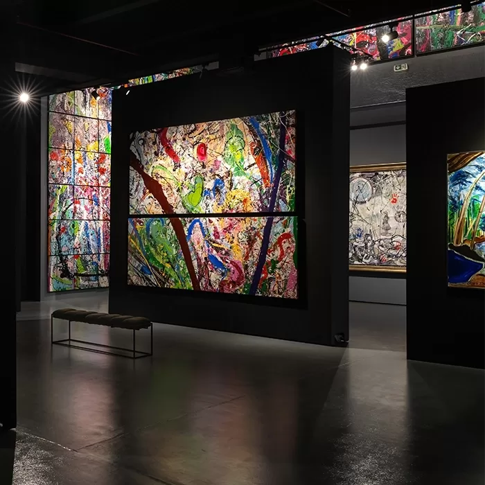 Large, colorful abstract paintings with bold, swirling patterns are displayed on dark walls in a modern art gallery. The polished concrete floor and spotlights create a dramatic and elegant atmosphere. A simple bench sits nearby.