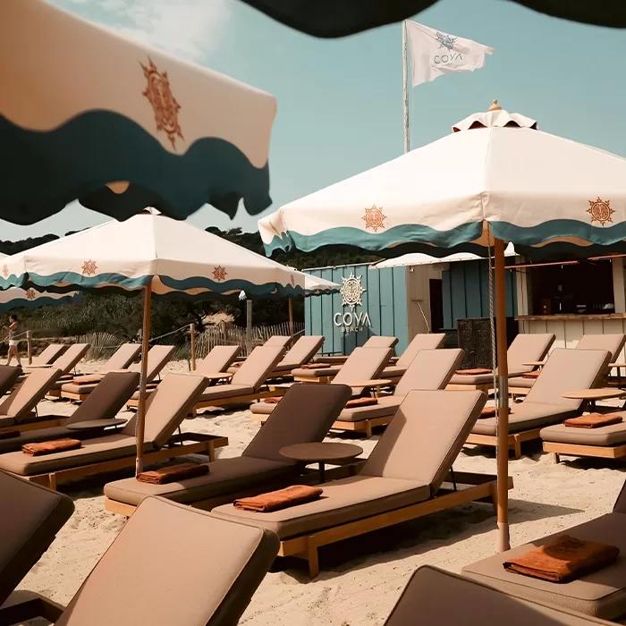 Rows of cushioned lounge chairs with brown towels sit under white and blue sun umbrellas on a sandy beach. A flag and a small beach bar with COVA signage are visible in the background under a sunny sky.