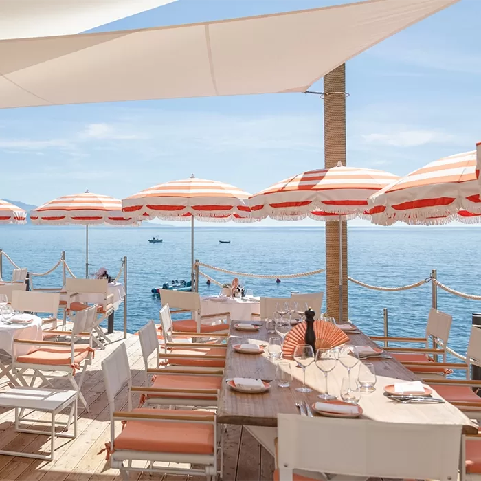 Outdoor seaside dining area with white and orange striped umbrellas, white chairs, and set tables overlooking calm blue water, with boats and people swimming in the background under a sunny sky.