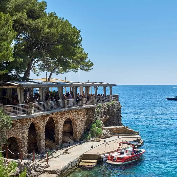 A seaside restaurant with arched stone walls sits on a rocky terrace above clear blue water, shaded by tall trees. A small red and white boat is docked at the pier below under a sunny sky.
