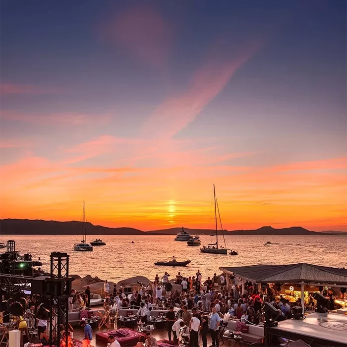 A vibrant beach party at sunset with people socializing, dining, and relaxing. Sailboats and yachts are anchored on calm water under an orange and pink sky. The scene is lively and festive with scenic coastal views.